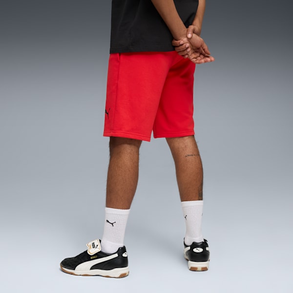 Against grey, a model stands wearing red shorts, a black t-shirt, shoes and white socks.