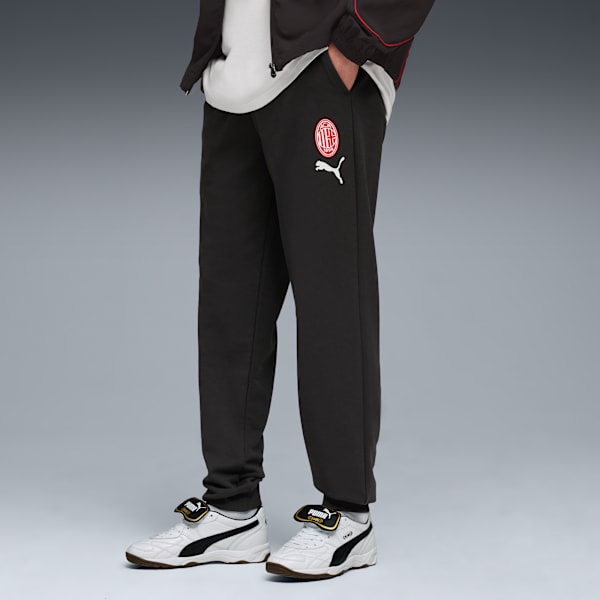 Against a grey backdrop, a model poses in black joggers and white trainers.