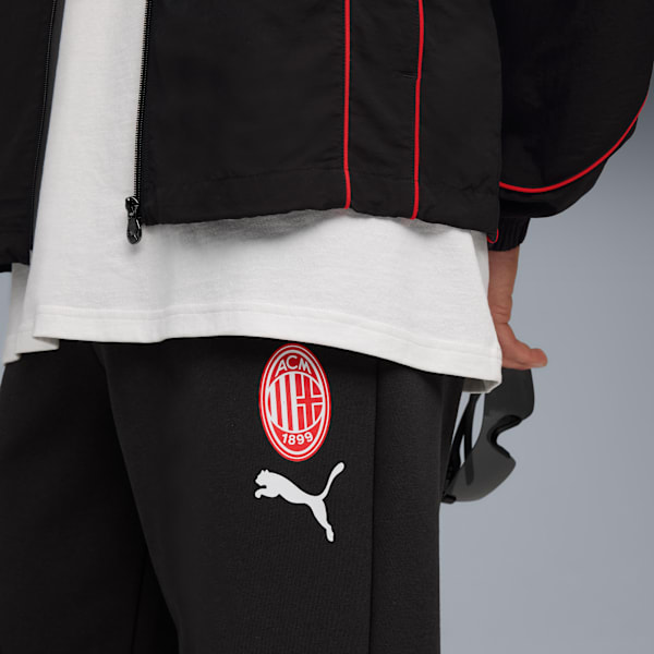 A model wears a black jacket, white t-shirt and black trousers with logos in front of a grey background.