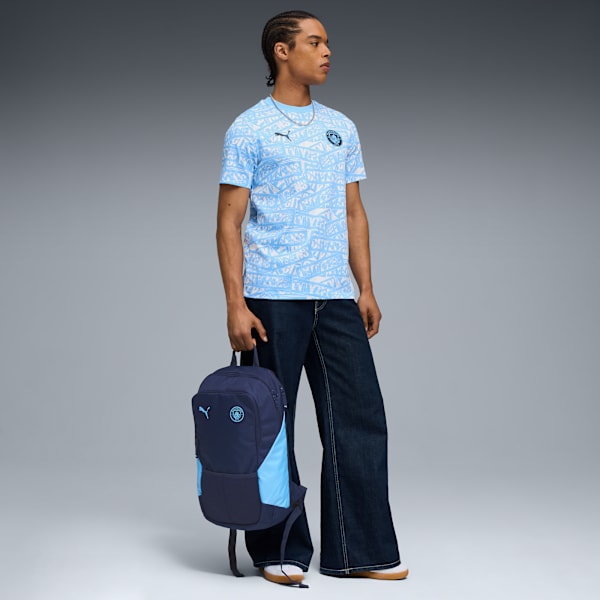 On grey, a model wearing jeans and a patterned shirt holds a two-tone navy and light blue rucksack.