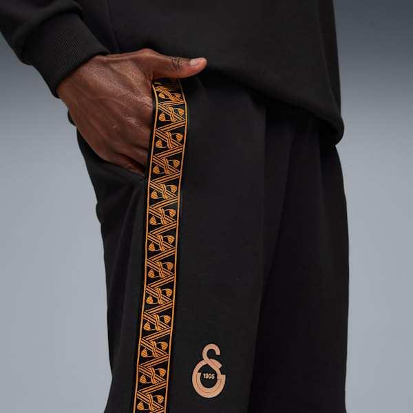 Against grey, a person wears black trousers with patterned side tape and hand in pocket.