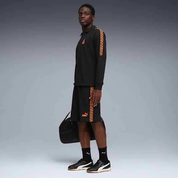 Against grey, a model poses in black shirt, shorts, trainers, holding a bag.
