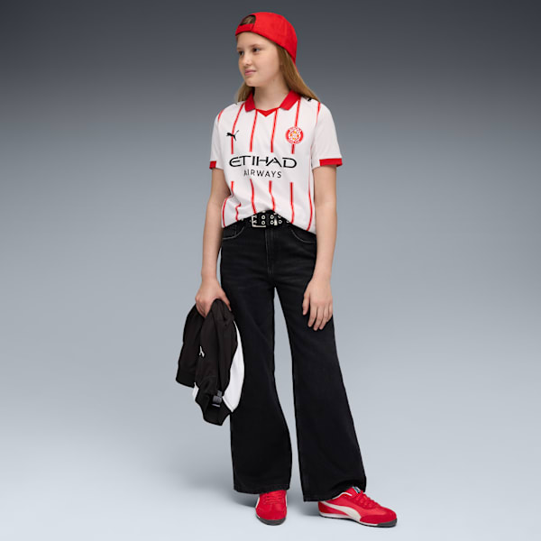 Against grey, a model poses in jeans, red shoes, and a striped t-shirt, holding a jacket.