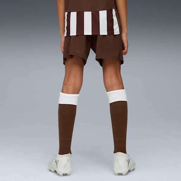 On grey, a model wears brown shorts, shirt with stripes, brown socks and trainers.