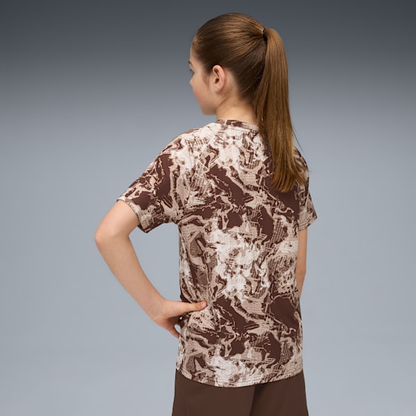 A model with ponytail, seen from behind, wears a brown patterned t-shirt, against grey.