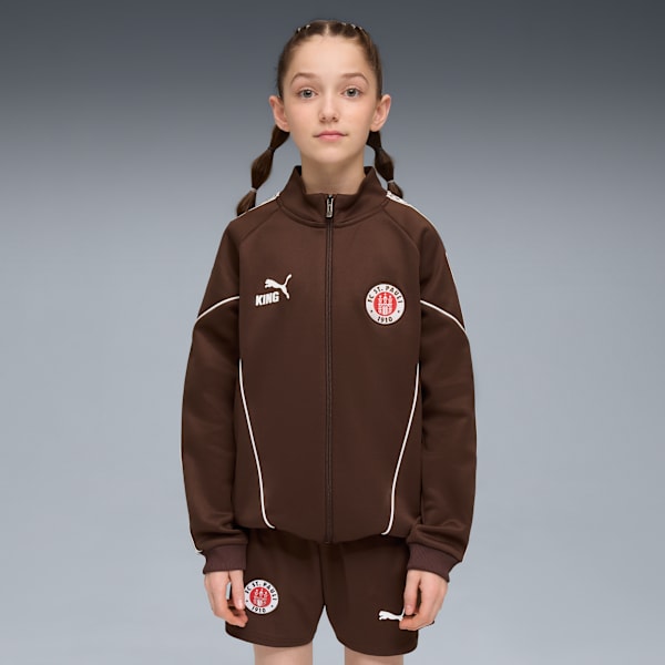 Against grey, a model poses in a brown jacket and shorts with team emblems.