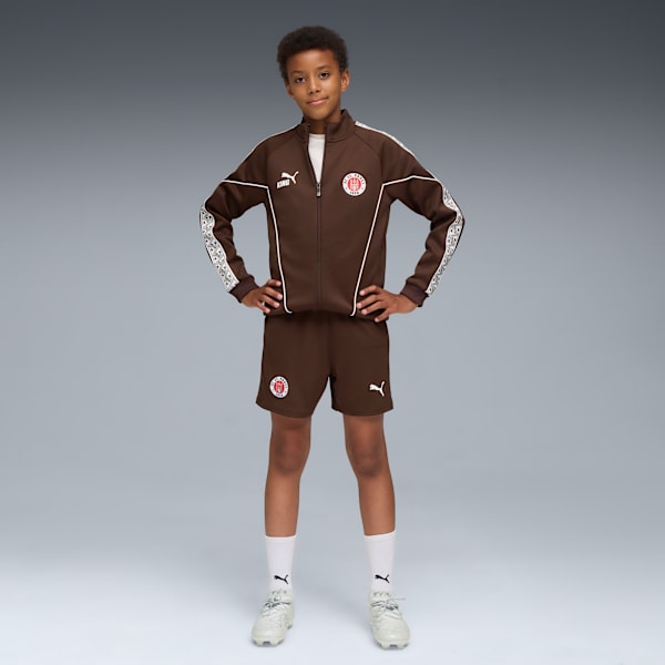 A model stands against a grey backdrop in a brown jacket and shorts with white socks.