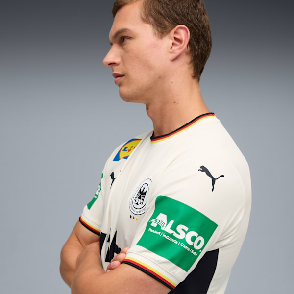 Against grey, a model stands with arms crossed, wearing a white t-shirt with German flag colours.