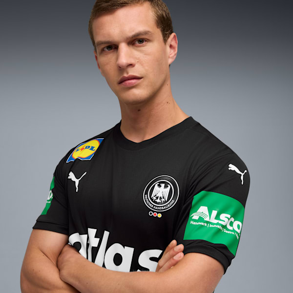 A model with crossed arms wears a black t-shirt with sponsor logos against a grey backdrop.