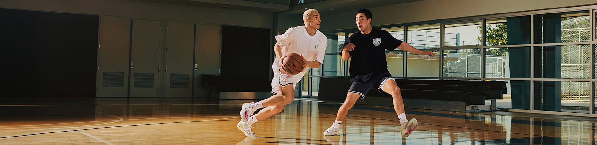Two people in sportswear play basketball on a court with large windows.