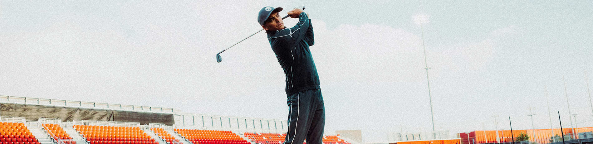 Against stadium seating, a golfer in navy sportswear swings their club.