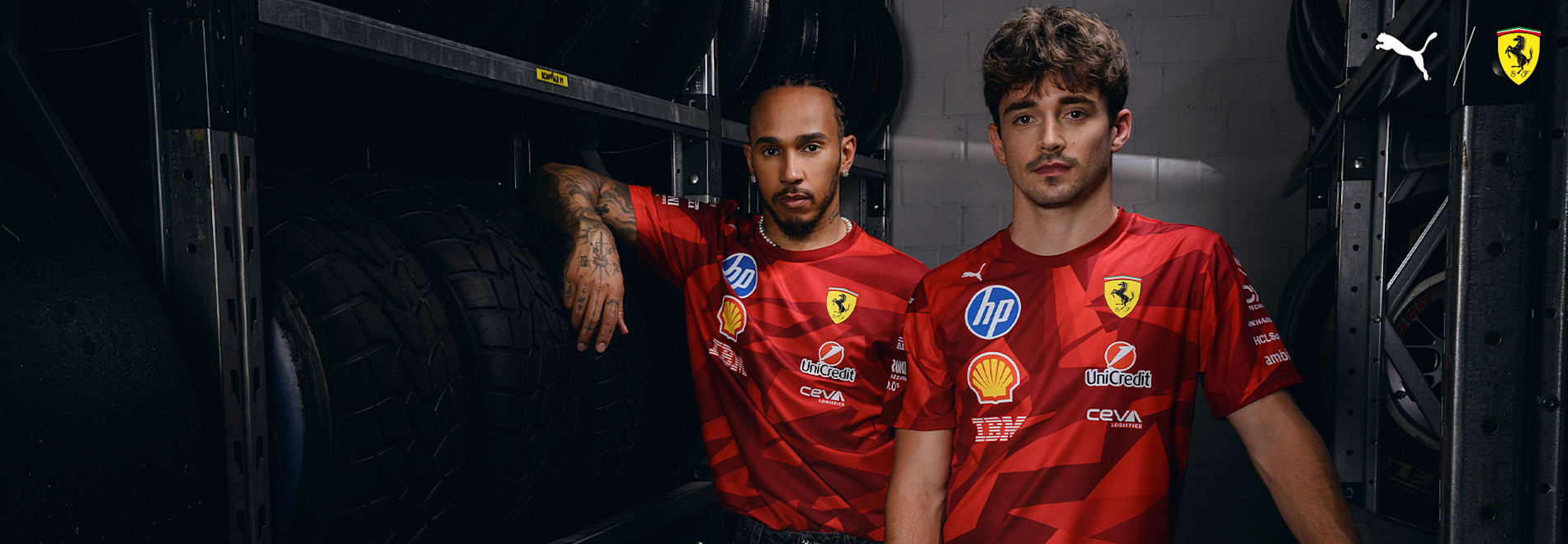 Two models in red Ferrari shirts pose by stacked tyres in a garage setting.