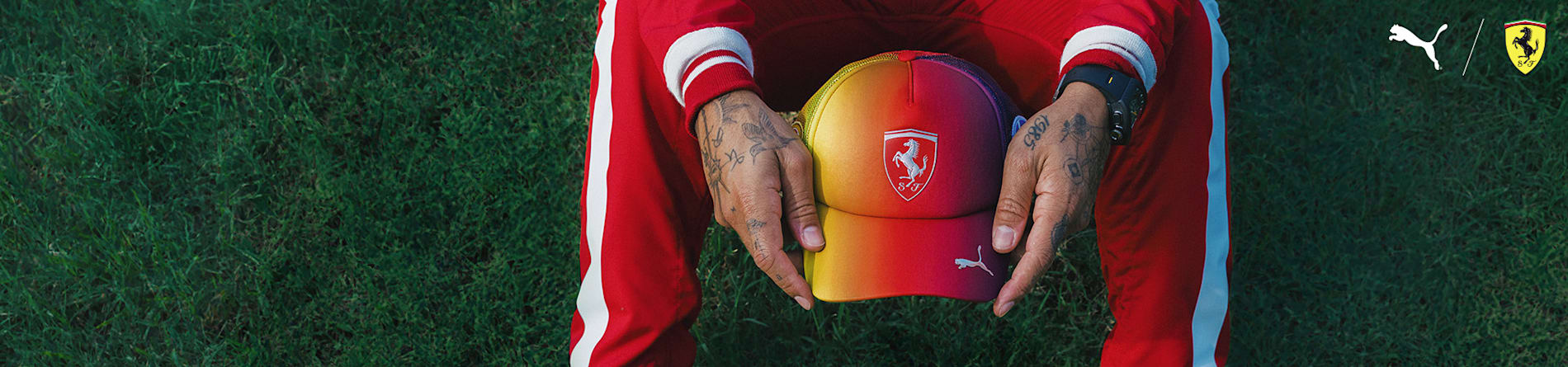 On grass, tattooed hands hold a Ferrari cap with red-yellow fade, matching red-white tracksuit.