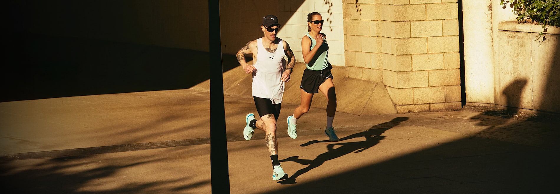 Athletes in running gear and light teal trainers run past a stone wall in bright sunlight.