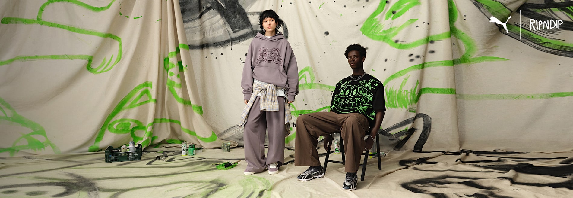 Two models pose in Puma x Ripndip apparel against a painted backdrop, one seated.