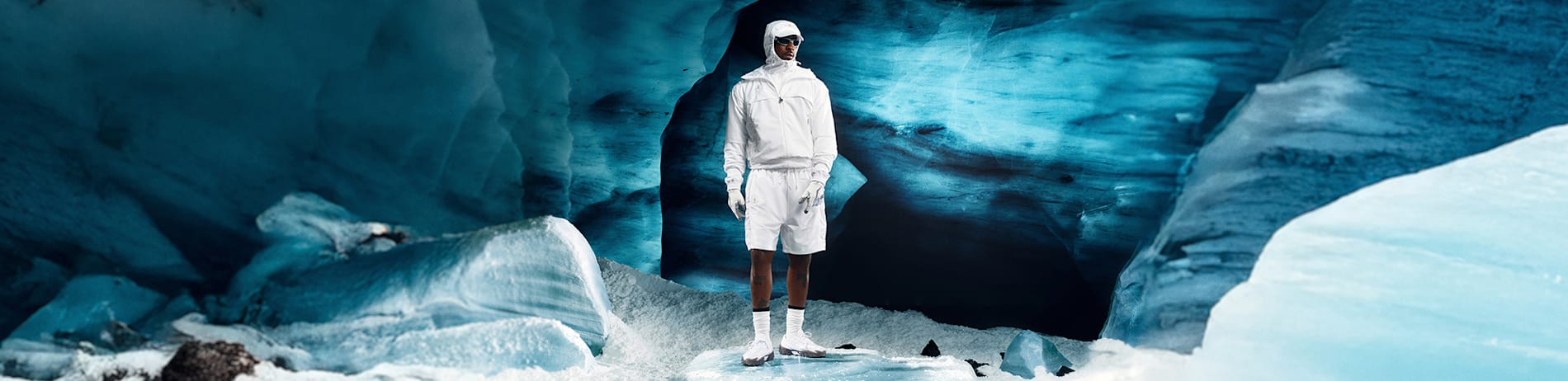 Against a blue ice cave stands a model in white sportswear with sunglasses.