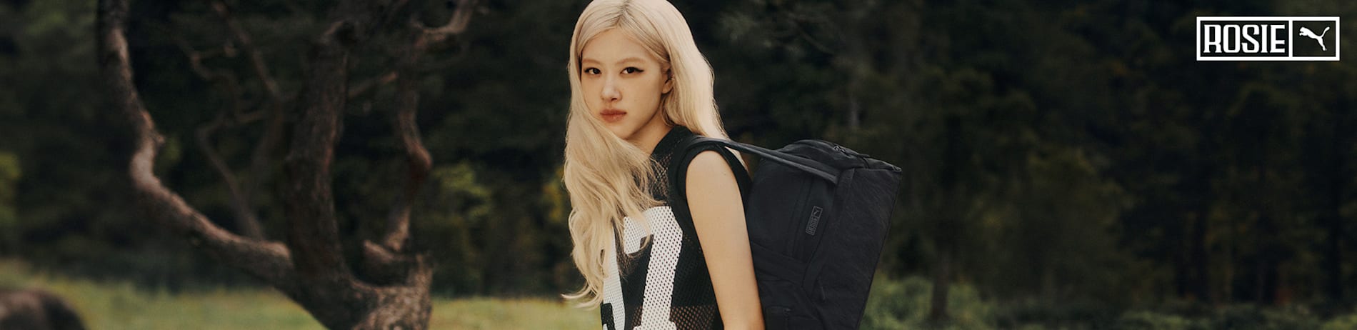Blonde model in sporty black outfit with bag poses in a blurred forest background.
