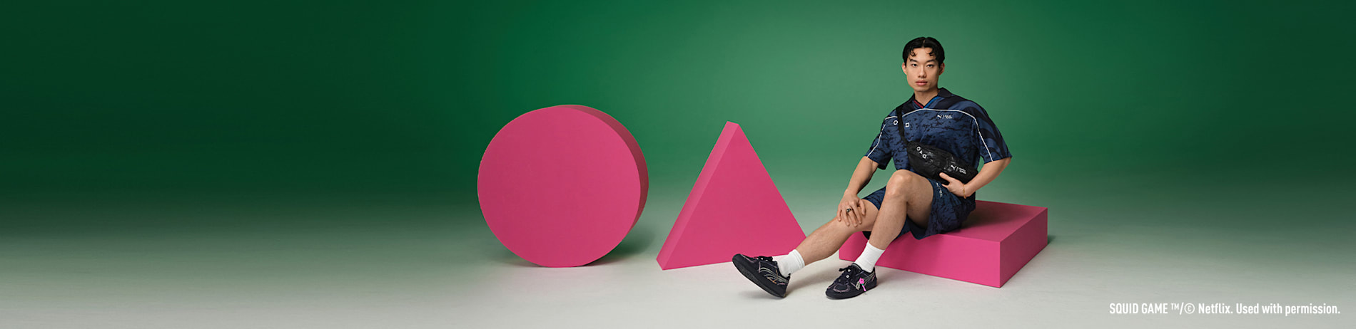 Against green, a model in Squid Game-themed outfit sits by pink shapes, wearing branded trainers.