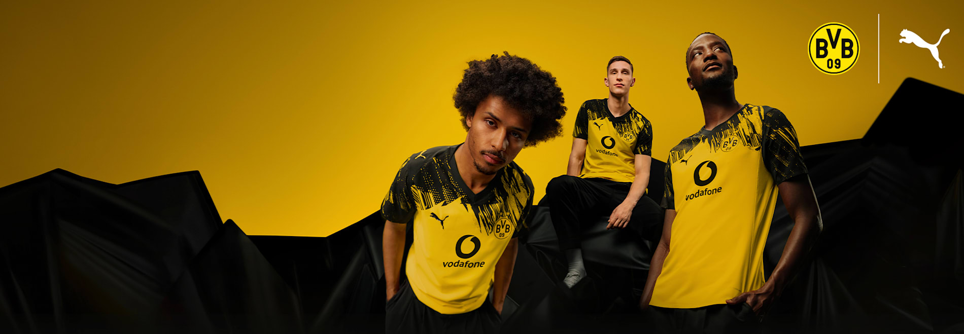 Three models pose in yellow and black Borussia Dortmund football shirts against a yellow backdrop.