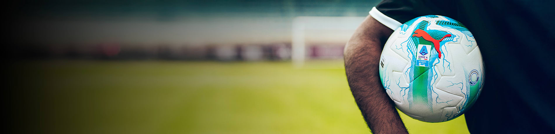 A person holds a white Serie A football with blue accents and a red logo against a blurred field backdrop.