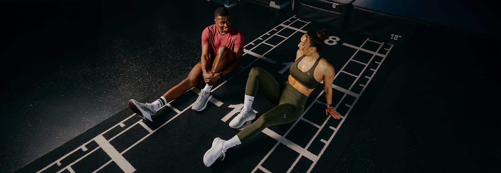 En un gimnasio, dos modelos en ropa deportiva se sientan en un suelo marcado, sonriendo y descansando.