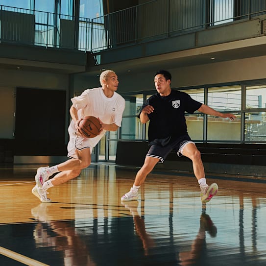 Zwei Personen in Sportkleidung spielen Basketball auf einem Platz mit grossen Fenstern.