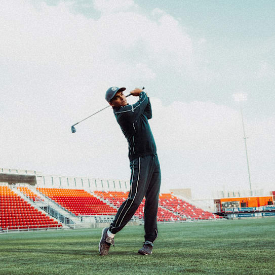 A golfer in sportswear swings a club on a green field with stadium seating behind.