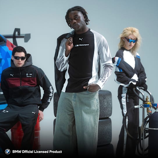 Drei Models posieren in BMW Motorsport-Bekleidung mit Reifen und einem Rennwagenflügel in der Nähe.