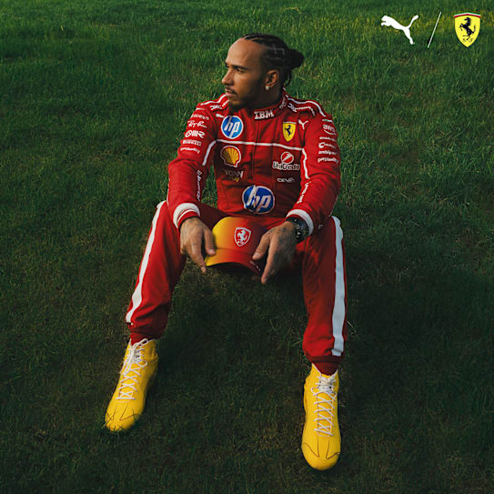 Sur l'herbe, une personne en combinaison Ferrari rouge et chaussures jaunes tient une casquette.