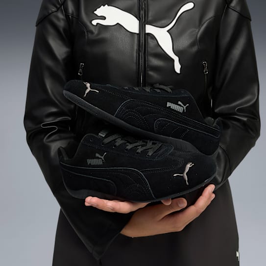 A person in a jacket with a logo holds two black Puma trainers against a grey background.