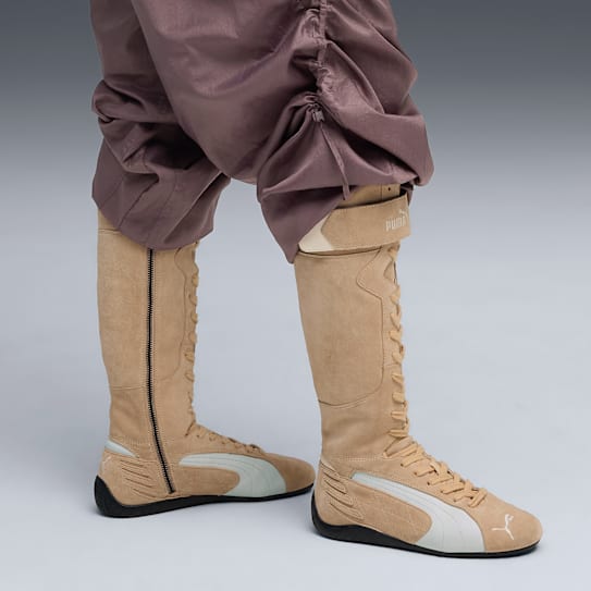 On grey, a model wears tan, laced, knee-high boots with the Puma logo and mauve gathered trousers.