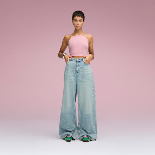 Sur un fond rose, un mannequin pose en haut rose, jean large et chaussures vertes.