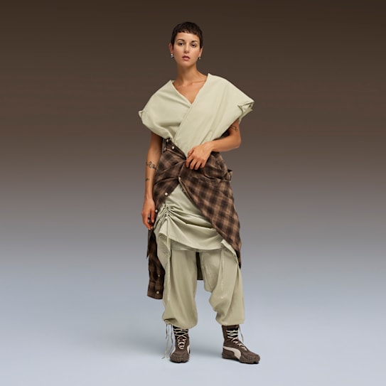 Against a gradient backdrop, a model poses in a draped light green outfit and brown trainers.