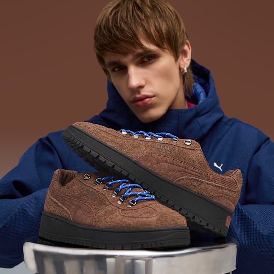 A model in a blue hooded jacket holds brown Palermo trainers with blue laces.