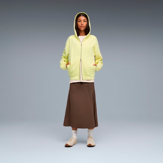 On grey, a model poses in a yellow hooded jacket, brown skirt, socks and trainers.