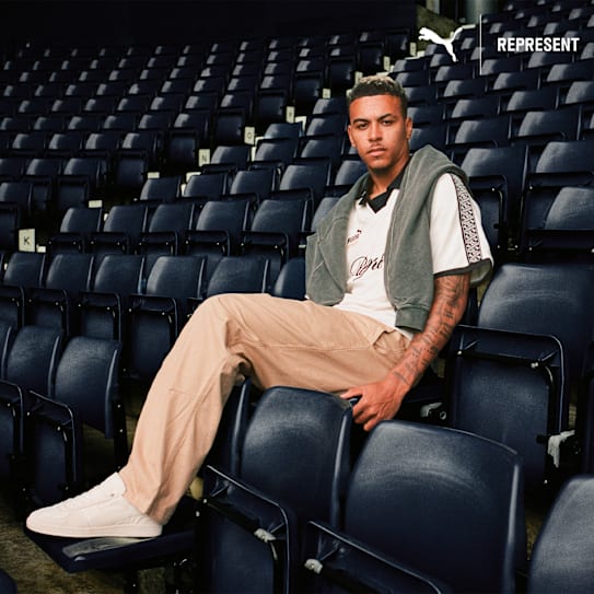 Eine Person sitzt in einem Stadion auf einem Sitzplatz und trägt Turnschuhe, Hose und ein Puma-Shirt.