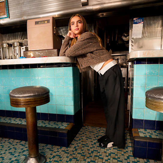 In a diner, a child model leans on the counter wearing black trainers, trousers, and a cardigan.