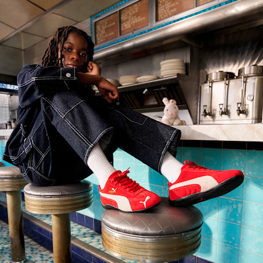 In a diner, a child sits on a stool wearing red trainers and a denim outfit.