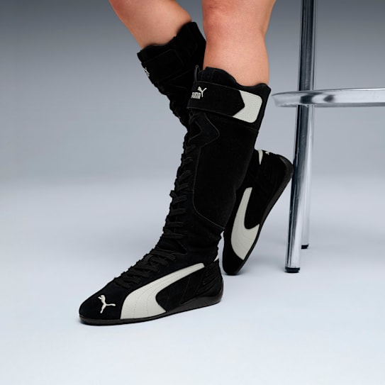 On grey, legs wear black, lace-up Puma boots with white stripes and adjustable straps.