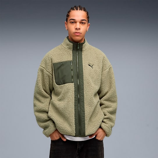 Vor grauem Hintergrund posiert ein Mann in einer grünen Fleecejacke mit Reißverschluss und dunklen Jeans.