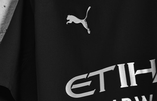 Close-up van een zwart voetbalshirt met een zilveren logo en sponsorprint tegen een grijze achtergrond.