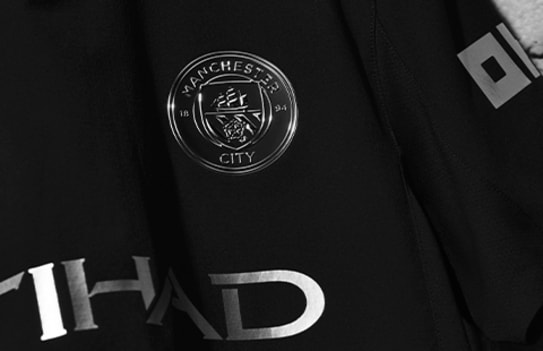 Close-up van een zwart Manchester City uitshirt met zilveren logo details.