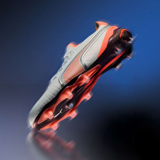 Une chaussure de foot grise et orange est suspendue sur un fond bleu flou.