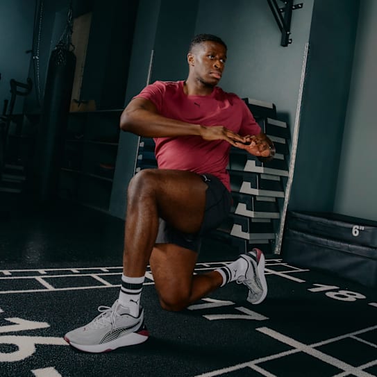 In einem Fitnessstudio wärmt sich ein Mann in Sportkleidung mit Kniebeugen auf, trägt Turnschuhe und Socken.