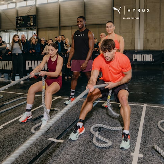 In a gym, models in sportswear pull ropes during a Hyrox training event, watched by a crowd.