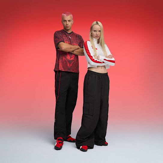 Two models pose in red, black and white outfits with arms crossed, against a red backdrop.
